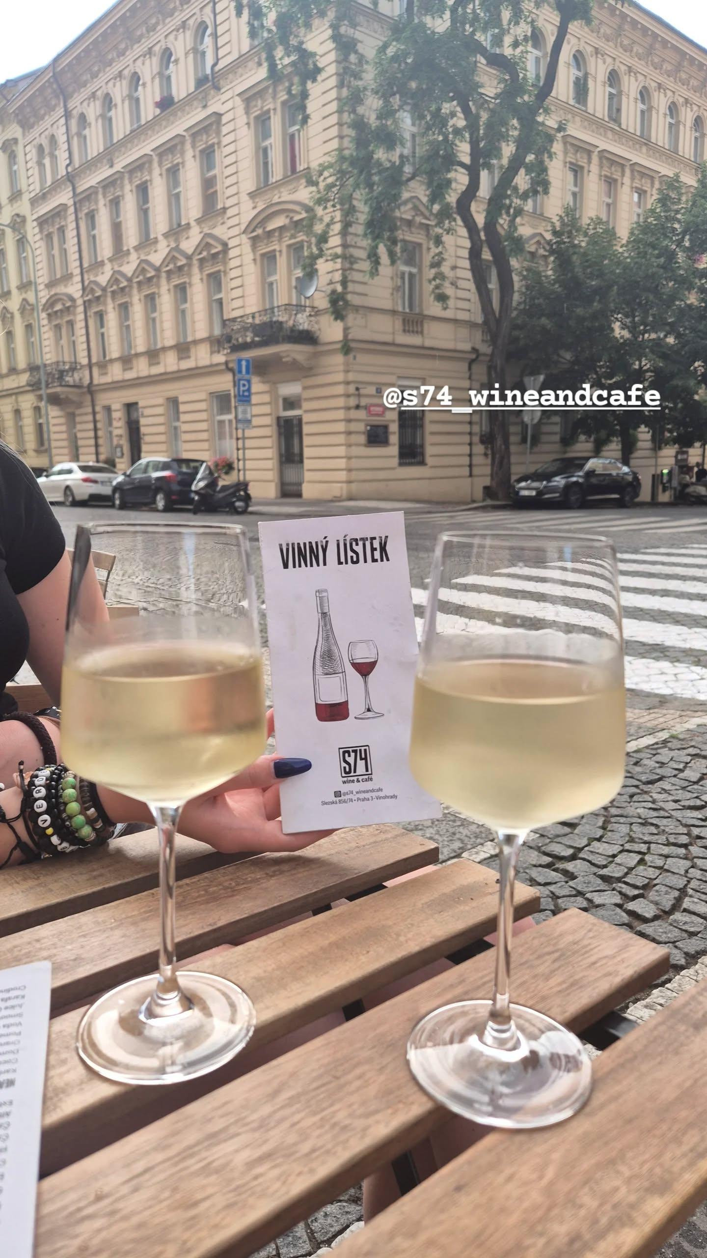 Instagramové story z S74 wine&café: dvě sklenice bílého vína na stole na terase a v ruce vinný lístek, na rohu pražské ulice